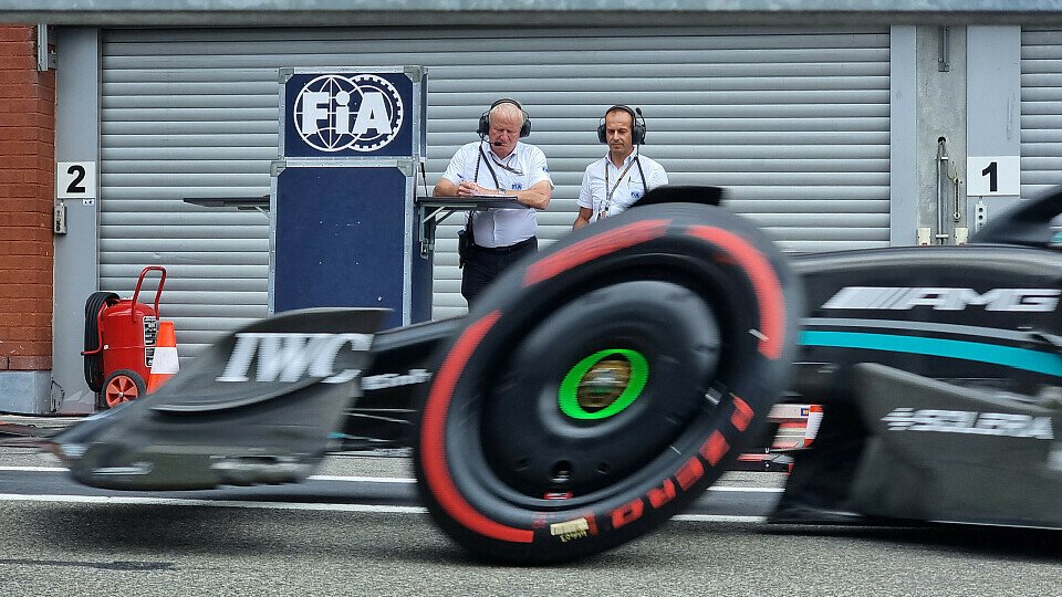 Zwei Angestellte der FIA in der Formel-1-Boxengasse werden von einem Mercedes passiert.